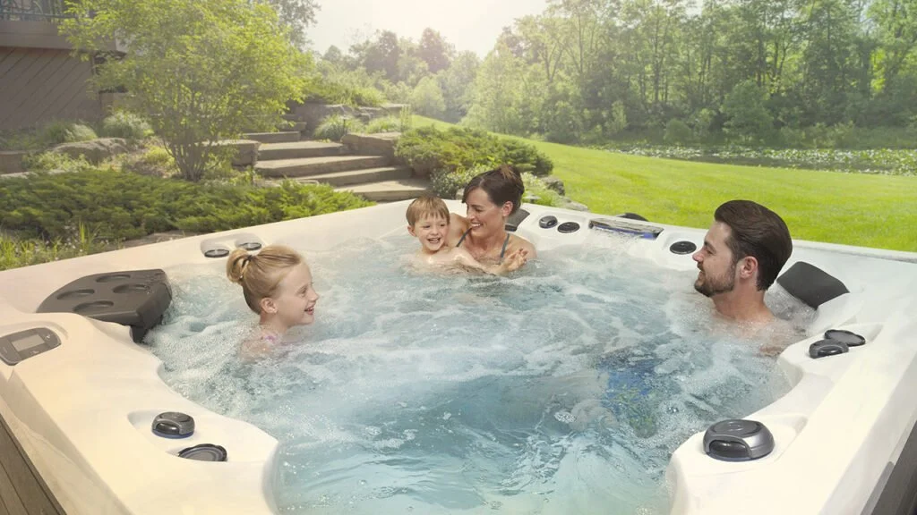 Family sitting together in a backyard hot tub during daytime, surrounded by grass, trees, and garden space